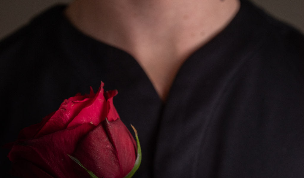 Close up on the neck of a man wearing a Cheegs collarless dress shirt in black holding a rose.