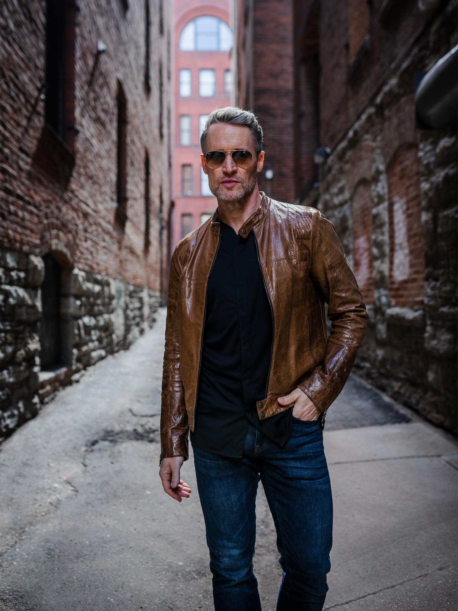 A young man wearing black dress shirt that is collarless but formal. Collarless shirt is styled with jeans and a brown leather jacket