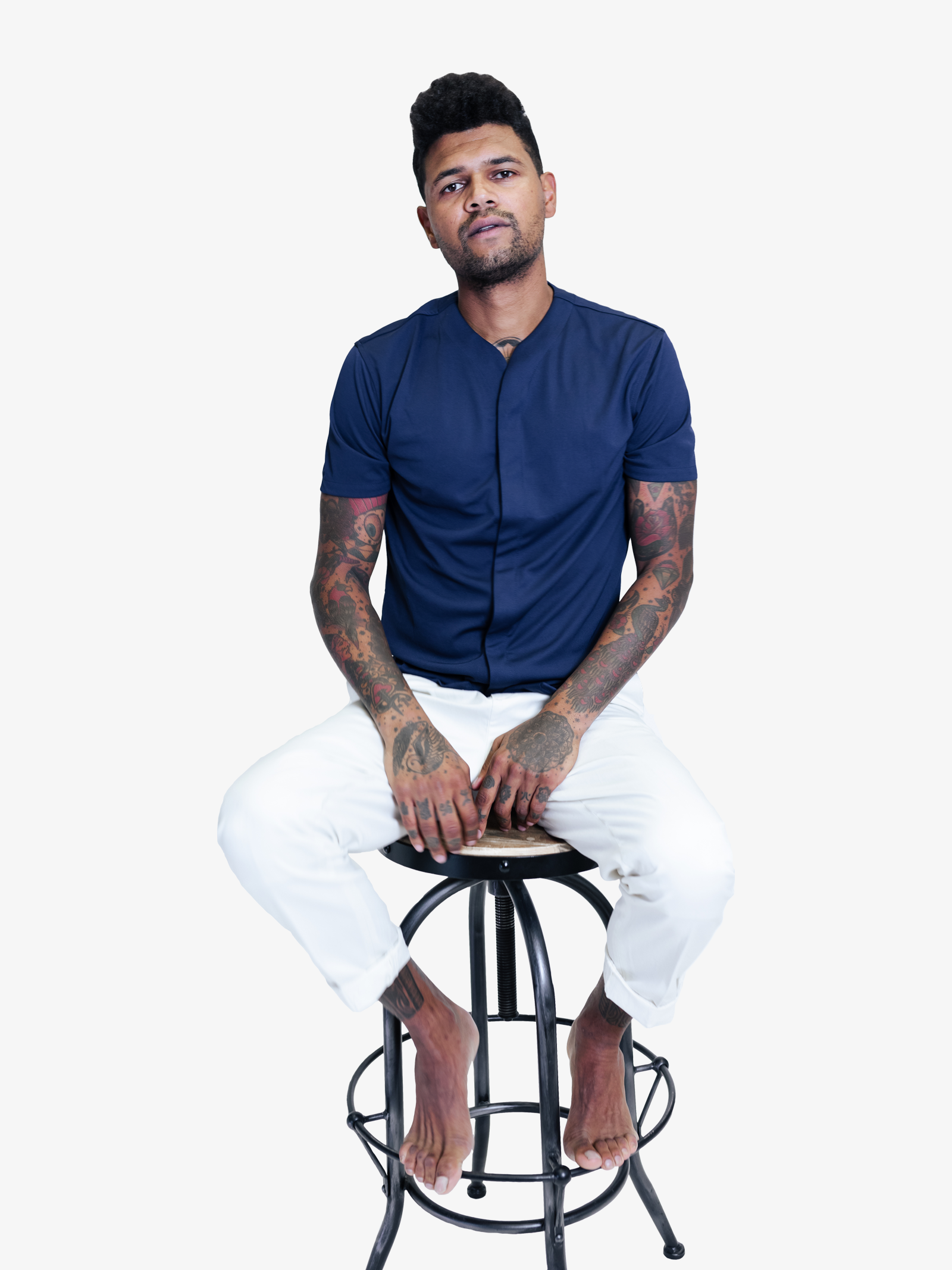 Model with tattoos on a stool wearing a collarless short sleeve shirt in blue and white pants